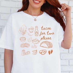 May include: White t-shirt featuring a brown illustration of taco ingredients and the text "tacos for two please". The graphic includes images of lettuce, salsa, a taco, and other food items. The shirt has a relaxed, oversized fit.