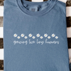 May include: A blue t-shirt with the text "growing two tiny humans" in white script, adorned with a row of white daisy flowers. The shirt is folded and placed on a black and white checkered surface, with a pair of blue jeans and a hat in the background.