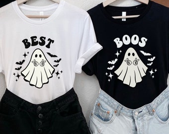 May include: Two white t-shirts with a ghost graphic and the text "BEST" and "BOOS" printed on them. The shirts are displayed on hangers with a brown hat on each side.