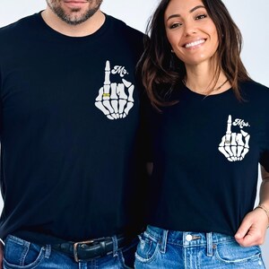 Pode incluir: Um casal vestindo camisetas pretas combinando com um design de m&atilde;o de esqueleto e as palavras "Mr." e "Mrs." impressas nelas. A m&atilde;o de esqueleto est&aacute; usando um anel no dedo indicador.