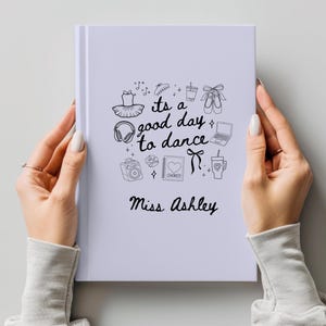 May include: A white notebook with the text "its a good day to dance" and the name "Miss Ashley" in black script. The cover features black line drawings of ballet-related items. The notebook is held by two hands.