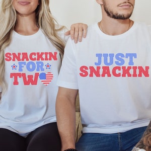 May include: A couple wearing white t-shirts with red, white and blue text. The woman's shirt says "Snackin' for Two" with a small American flag heart. The man's shirt says "Just Snackin'" in red and blue text.