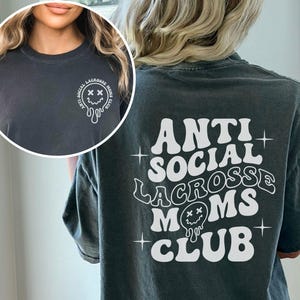 May include: A dark gray t-shirt with a white graphic that says "Anti Social Lacrosse Moms Club" in a retro font. The graphic includes a dripping smiley face with X's for eyes.