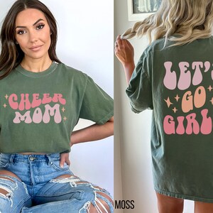 May include: Two green t-shirts with a retro font design. One shirt says "Cheer Mom" and the other says "Let's Go Girls".