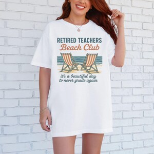 May include: White t-shirt with the text "Retired Teachers Beach Club" and a graphic of two beach chairs. The text below the graphic reads "It's a beautiful day to never grade again."