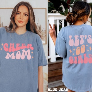 May include: A light blue t-shirt with the text "Cheer Mom" in pink and orange retro font. The back of the shirt has the text "Let's Go Girls" in pink and orange retro font.