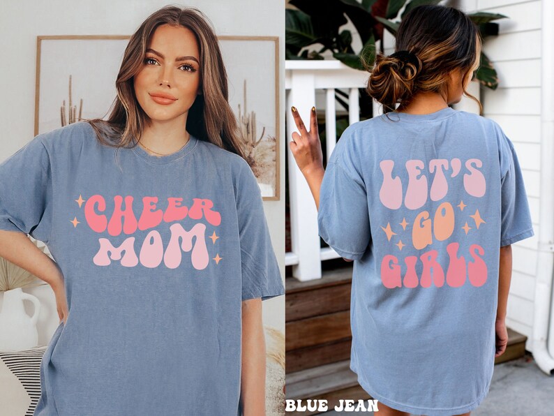 May include: A light blue t-shirt with the text "Cheer Mom" in pink and orange retro font. The back of the shirt has the text "Let's Go Girls" in pink and orange retro font.