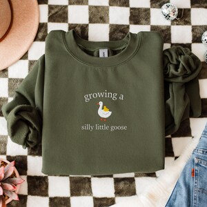 K&ouml;nnte beinhalten: Ein olivgr&uuml;nes Sweatshirt mit dem Schriftzug "growing a silly little goose" und einer Grafik einer wei&szlig;en Gans und eines gelben K&uuml;ken. Das Sweatshirt liegt gefaltet auf einer karierten Oberfl&auml;che, mit einem rosa Hut und Jeans sichtbar.