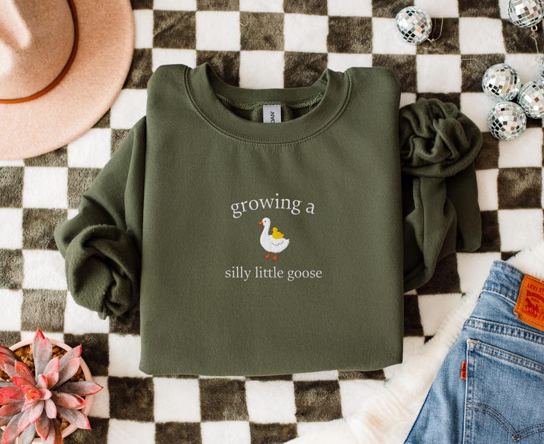 K&ouml;nnte beinhalten: Ein olivgr&uuml;nes Sweatshirt mit dem Schriftzug "growing a silly little goose" und einer Grafik einer wei&szlig;en Gans und eines gelben K&uuml;ken. Das Sweatshirt liegt gefaltet auf einer karierten Oberfl&auml;che, mit einem rosa Hut und Jeans sichtbar.