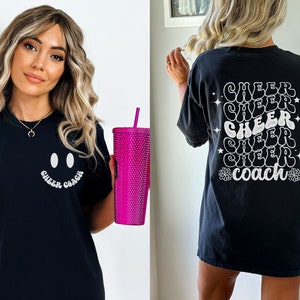 Peut inclure: T-shirt noir avec un smiley blanc et le texte "Cheer Coach" sur le devant. Le dos du t-shirt porte le texte "Cheer" r&eacute;p&eacute;t&eacute; cinq fois dans une police r&eacute;tro, avec le mot "Coach" au centre.
