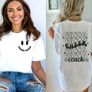 Peut inclure: T-shirt blanc avec un smiley noir et le texte "Cheer Coach" sur le devant. Le dos du t-shirt porte le texte "Cheer" r&eacute;p&eacute;t&eacute; cinq fois dans une police r&eacute;tro, suivi de "Coach" avec une fleur de chaque c&ocirc;t&eacute;.