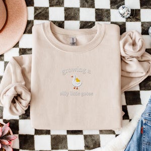 K&ouml;nnte beinhalten: Beiges Sweatshirt mit dem gestickten Schriftzug "growing a silly little goose" und einer Cartoon-Gans. Das Sweatshirt liegt auf einer schwarz-wei&szlig; karierten Oberfl&auml;che, mit einem rosa Hut und Jeans im Hintergrund.