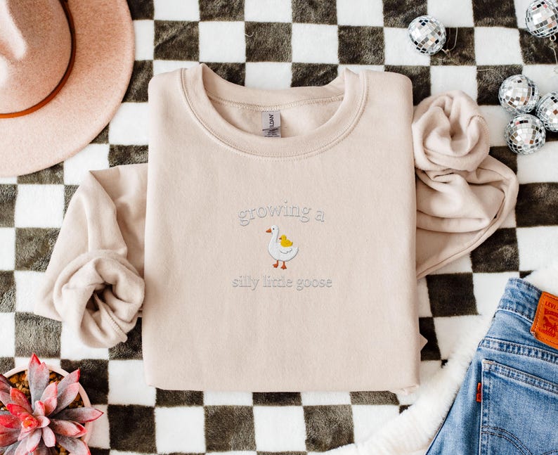 K&ouml;nnte beinhalten: Beiges Sweatshirt mit dem gestickten Schriftzug "growing a silly little goose" und einer Cartoon-Gans. Das Sweatshirt liegt auf einer schwarz-wei&szlig; karierten Oberfl&auml;che, mit einem rosa Hut und Jeans im Hintergrund.