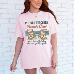 May include: Pink t-shirt with the text "Retired Teachers Beach Club" and a graphic of two beach chairs. The text below the graphic reads "It's a beautiful day to never grade again."