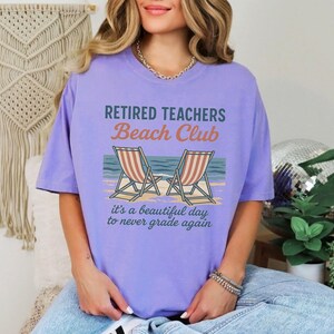 May include: Purple t-shirt with the text "Retired Teachers Beach Club" and a graphic of two beach chairs. The text below the graphic reads "it's a beautiful day to never grade again."