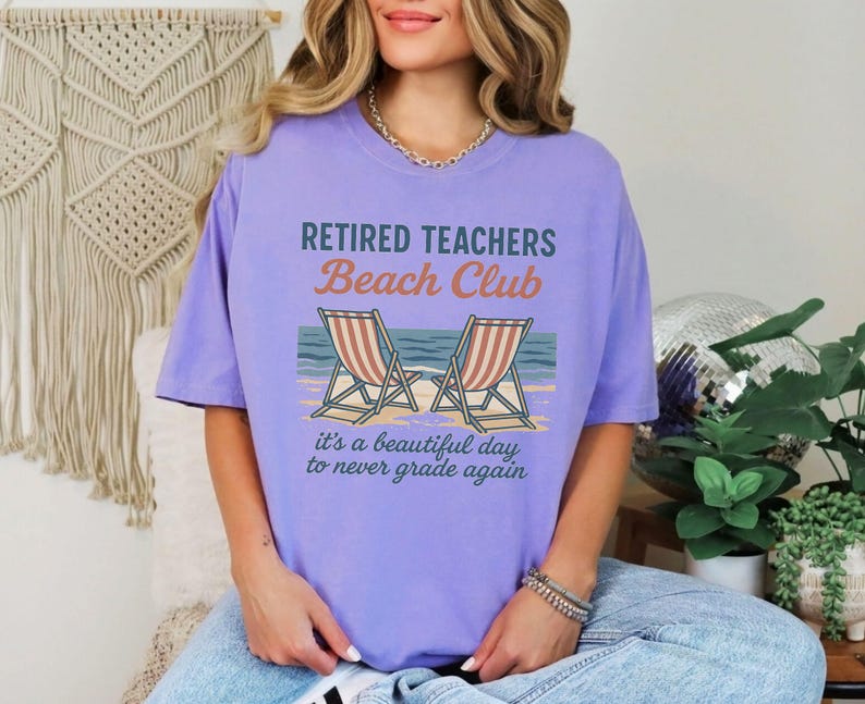 May include: Purple t-shirt with the text "Retired Teachers Beach Club" and a graphic of two beach chairs. The text below the graphic reads "it's a beautiful day to never grade again."