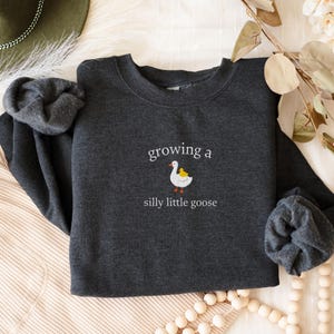 K&ouml;nnte beinhalten: Dunkelgraues Sweatshirt mit dem Text "growing a silly little goose" und einer G&auml;nse-Cartoon-Grafik. Die Gans ist wei&szlig; mit gelbem L&auml;tzchen und orangefarbenen F&uuml;&szlig;en. Das Sweatshirt ist auf einer cremefarbenen Oberfl&auml;che gefaltet.