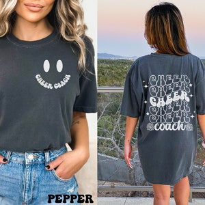 Peut inclure: Deux femmes portant des t-shirts gris fonc&eacute; avec du texte blanc. Le t-shirt de gauche a un smiley et le texte "Cheer Coach". Le t-shirt de droite a le texte "Cheer" r&eacute;p&eacute;t&eacute; cinq fois et "Coach" en bas.