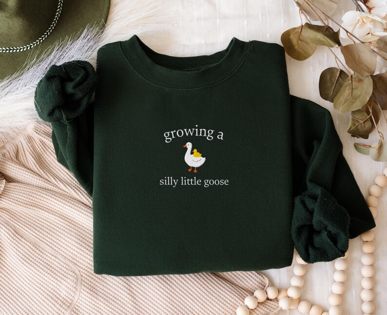 K&ouml;nnte beinhalten: Dunkelgr&uuml;nes Sweatshirt mit dem gestickten Schriftzug "growing a silly little goose" und einer Cartoon-Gans und einem Entlein. Das Sweatshirt hat lange &Auml;rmel und einen Rundhalsausschnitt.