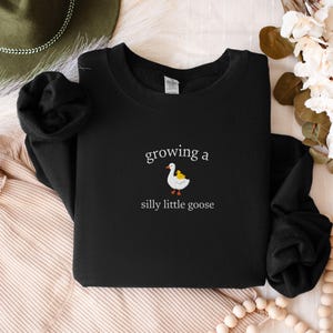 K&ouml;nnte beinhalten: Schwarzes Sweatshirt mit dem Text "growing a silly little goose" und einer Grafik einer wei&szlig;en Gans mit einem gelben K&uuml;ken. Das Sweatshirt hat einen Rundhalsausschnitt und ist aus weichem Material.