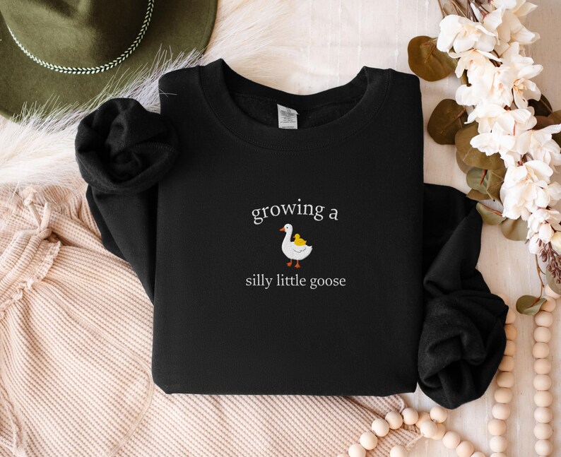 K&ouml;nnte beinhalten: Schwarzes Sweatshirt mit dem Text "growing a silly little goose" und einer Grafik einer wei&szlig;en Gans mit einem gelben K&uuml;ken. Das Sweatshirt hat einen Rundhalsausschnitt und ist aus weichem Material.