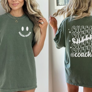 Peut inclure: Un t-shirt vert avec un smiley blanc et le texte "Cheer Coach" sur le devant. Le dos du t-shirt a le texte "Cheer Coach" r&eacute;p&eacute;t&eacute; cinq fois dans une police r&eacute;tro avec des &eacute;toiles et des fleurs.