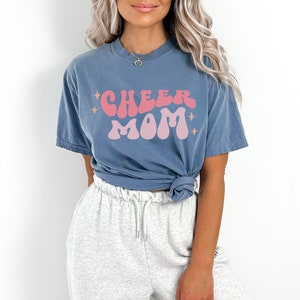 May include: A blue t-shirt with the text "CHEER MOM" in pink and peach retro lettering. The shirt is worn by a person with long blonde hair and grey sweatpants.