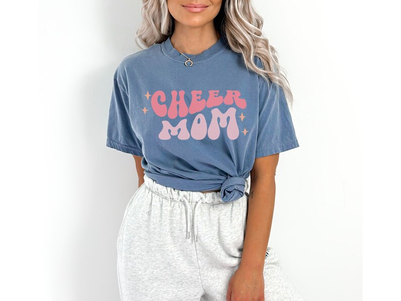 May include: A blue t-shirt with the text "CHEER MOM" in pink and peach retro lettering. The shirt is worn by a person with long blonde hair and grey sweatpants.