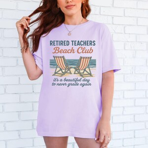 May include: A lavender t-shirt with the text "Retired Teachers Beach Club" and a graphic of two beach chairs with an ocean scene. The text below the graphic reads "It's a beautiful day to never grade again."