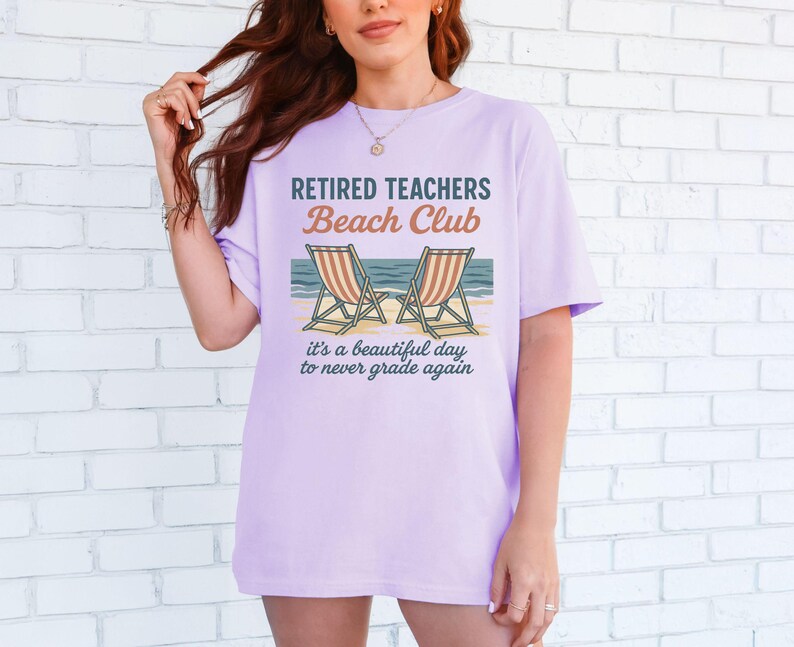 May include: A lavender t-shirt with the text "Retired Teachers Beach Club" and a graphic of two beach chairs with an ocean scene. The text below the graphic reads "It's a beautiful day to never grade again."