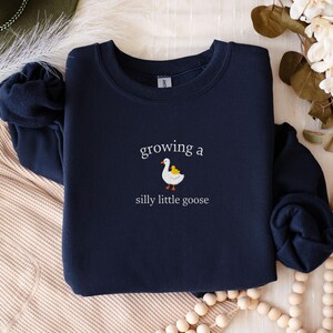 K&ouml;nnte beinhalten: Marineblaues Sweatshirt mit dem Text "growing a silly little goose" in wei&szlig;er Schrift. Unter dem Text ist eine Cartoon-Gans mit einem gelben K&uuml;ken abgebildet. Das Sweatshirt hat einen Rundhalsausschnitt.