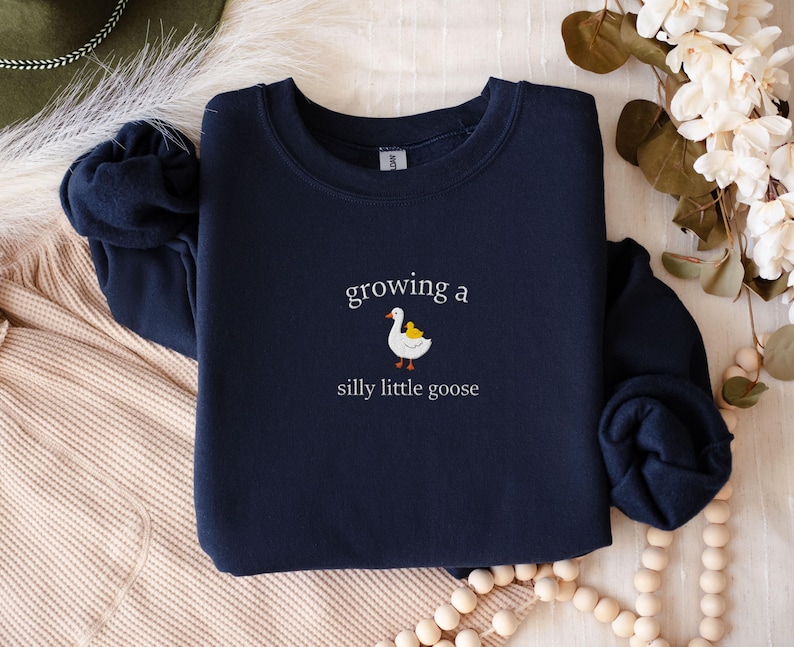 K&ouml;nnte beinhalten: Marineblaues Sweatshirt mit dem Text "growing a silly little goose" in wei&szlig;er Schrift. Unter dem Text ist eine Cartoon-Gans mit einem gelben K&uuml;ken abgebildet. Das Sweatshirt hat einen Rundhalsausschnitt.