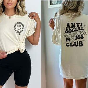 May include: A white t-shirt with a black graphic that says "Anti Social Lacrosse Moms Club" and a dripping smiley face with X's for eyes.
