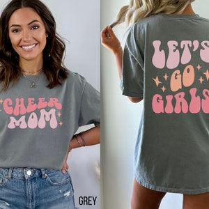 May include: A grey t-shirt with a pink and orange retro-style graphic that says "Cheer Mom" on the front and "Let's Go Girls" on the back.