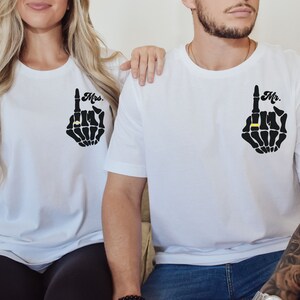 Pode incluir: Duas camisetas brancas com um design de m&atilde;o de esqueleto. A m&atilde;o aponta para cima com um anel no dedo indicador. Uma camiseta diz "Mr." e a outra "Mrs."