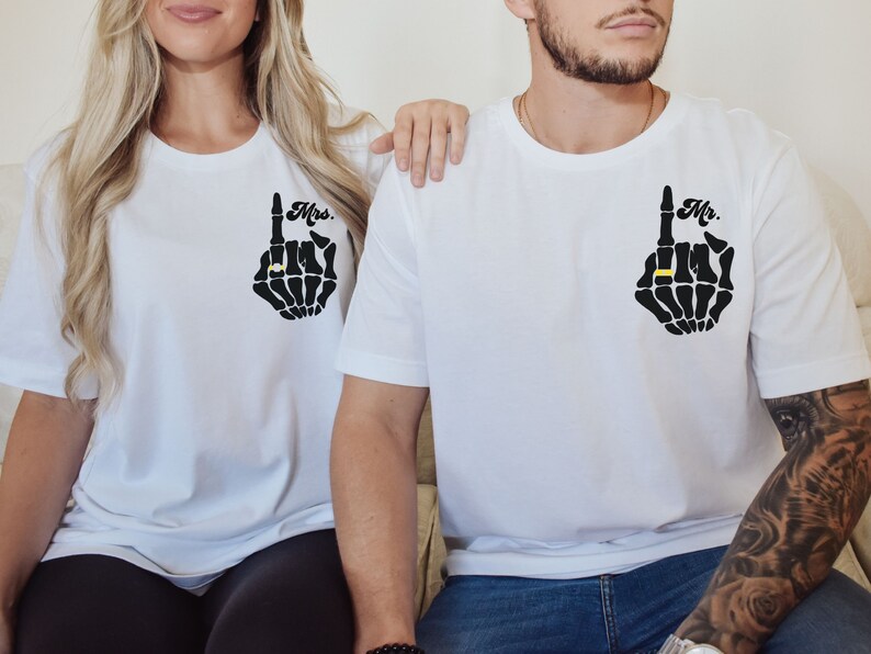 Pode incluir: Duas camisetas brancas com um design de m&atilde;o de esqueleto. A m&atilde;o aponta para cima com um anel no dedo indicador. Uma camiseta diz "Mr." e a outra "Mrs."