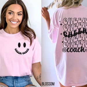 Peut inclure: T-shirt rose avec un smiley noir et le texte "Cheer Coach" sur le devant. Le dos du t-shirt a le texte "Cheer Coach" r&eacute;p&eacute;t&eacute; cinq fois dans une police r&eacute;tro.