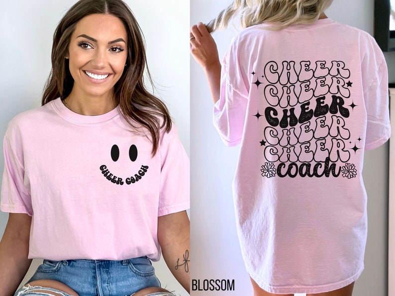 Peut inclure: T-shirt rose avec un smiley noir et le texte "Cheer Coach" sur le devant. Le dos du t-shirt a le texte "Cheer Coach" r&eacute;p&eacute;t&eacute; cinq fois dans une police r&eacute;tro.
