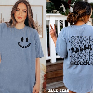 Peut inclure: Un t-shirt bleu avec un visage souriant et le texte "Cheer Coach" sur le devant. Le dos du t-shirt porte le texte "Cheer" r&eacute;p&eacute;t&eacute; cinq fois dans une police r&eacute;tro avec un motif de fleurs et d'&eacute;toiles.