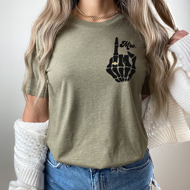 Pode incluir: Uma camiseta verde com uma m&atilde;o de esqueleto preta usando um anel e o texto "Mrs." impresso na frente.