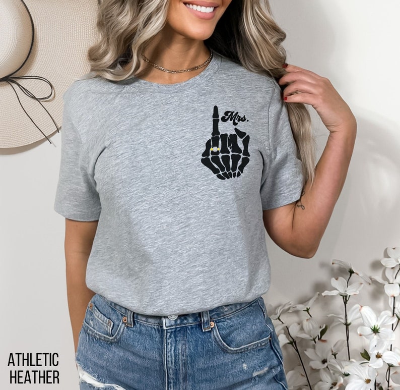 Pode incluir: Camiseta cinza com uma m&atilde;o de esqueleto preto usando um anel. A m&atilde;o est&aacute; fazendo um gesto de "um" e o texto "Mrs." est&aacute; acima da m&atilde;o.