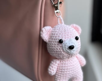 May include: A light pink crocheted bear keychain featuring a white muzzle, black plastic eyes, and a black nose. The bear is attached to a silver-toned metal lobster clasp and a short chain, allowing it to hang securely from a bag or accessory.