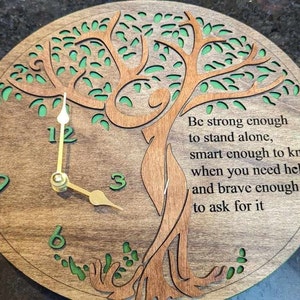 May include: A round wooden clock with a tree design and green accents. The clock face features the numbers 3, 6, 9, and 12, with gold-colored hands. The clock includes the text: "Be strong enough to stand alone..."