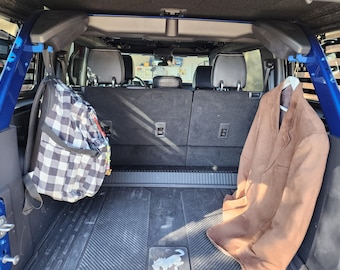 Könnte beinhalten: Der Rücksitz eines blauen SUV mit einem schwarz-weiß karierten Rucksack, der an einem Haken auf der linken Seite hängt, und einer braunen Jacke, die an einem Haken auf der rechten Seite hängt. Der Rücksitz ist leer und hat eine schwarze Gummimatte mit einem Logo.