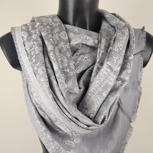 Può includere: Una sciarpa grigia con un motivo floreale bianco dettagliato e bordi sfrangiati è drappeggiata su un manichino. La sciarpa è realizzata in un materiale morbido e presenta un design a forma di paisley.