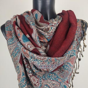 May include: A burgundy and paisley patterned scarf with teal, peach, and cream accents. The scarf is draped over a black mannequin and features a detailed paisley design. The scarf has a solid burgundy section and is finished with gold-colored fringe.
