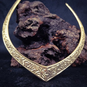 Puede incluir: Un collar gargantilla de metal dorado con un diseño floral. El collar se muestra sobre una roca marrón oscura.