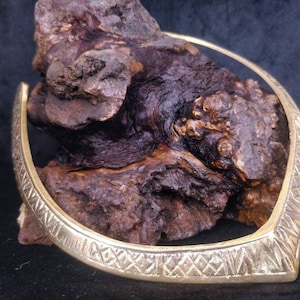 May include: A gold-coloured metal choker necklace with a geometric pattern. The necklace is displayed on a piece of dark brown wood.