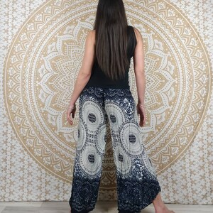 Puede incluir: Pantalones de pierna ancha con un estampado circular blanco y azul marino. Los pantalones tienen un borde azul marino oscuro en la parte inferior. La persona de la imagen lleva una camiseta sin mangas negra. El fondo es un tapiz mandala beige.