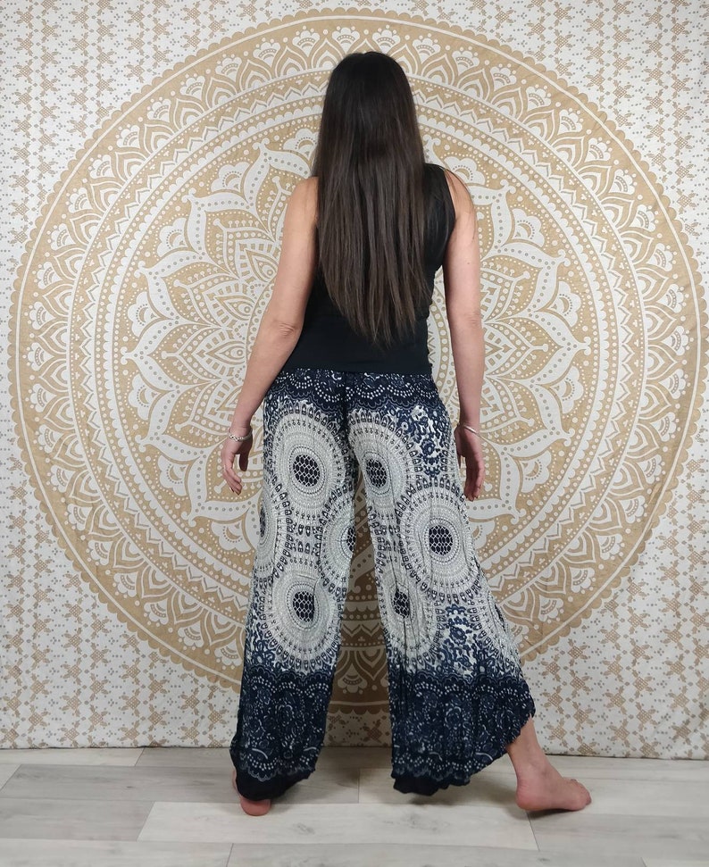 Puede incluir: Pantalones de pierna ancha con un estampado circular blanco y azul marino. Los pantalones tienen un borde azul marino oscuro en la parte inferior. La persona de la imagen lleva una camiseta sin mangas negra. El fondo es un tapiz mandala beige.